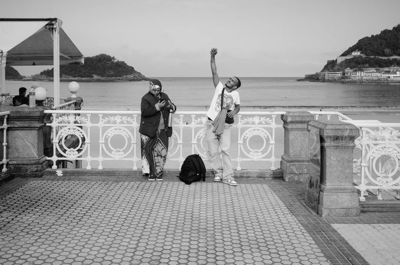 Two people at a white ironwork railing above the bay, one reaching a phone high over their head