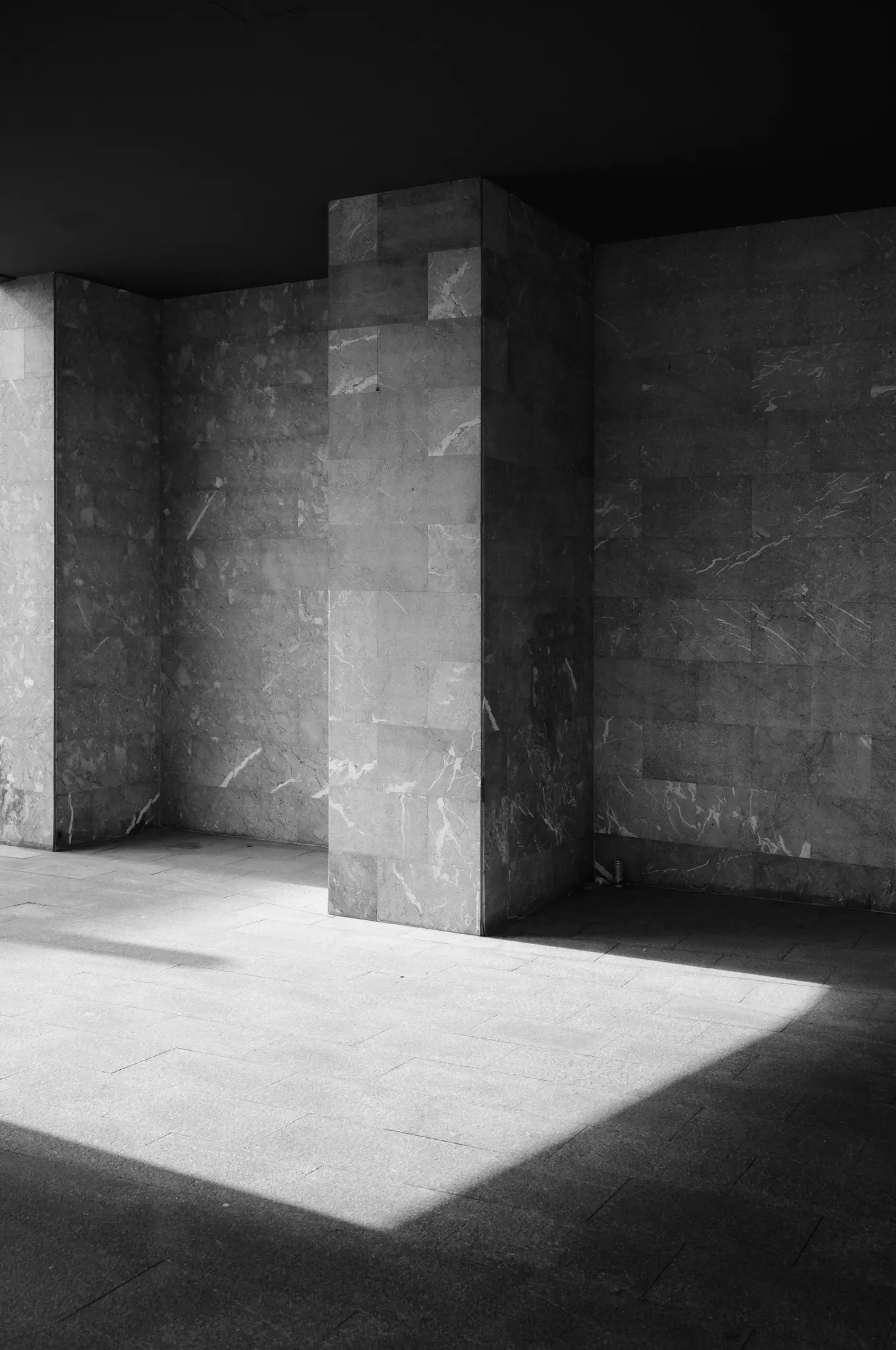 A square marble pillar in a shaded colonnade, sunlight cutting across the floor in front of it