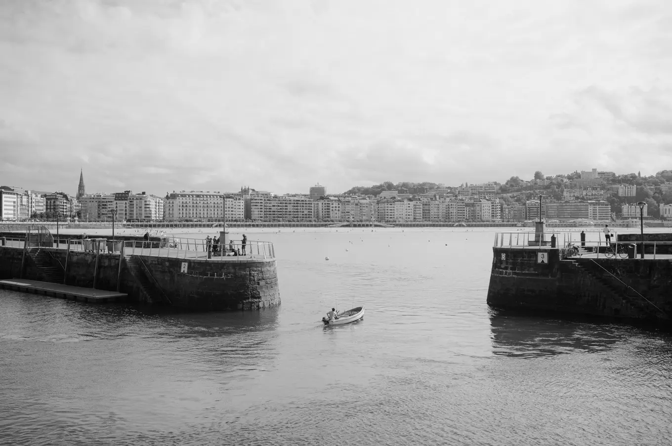 A small boat passing between two stone harbour piers, the seafront and its buildings across the bay