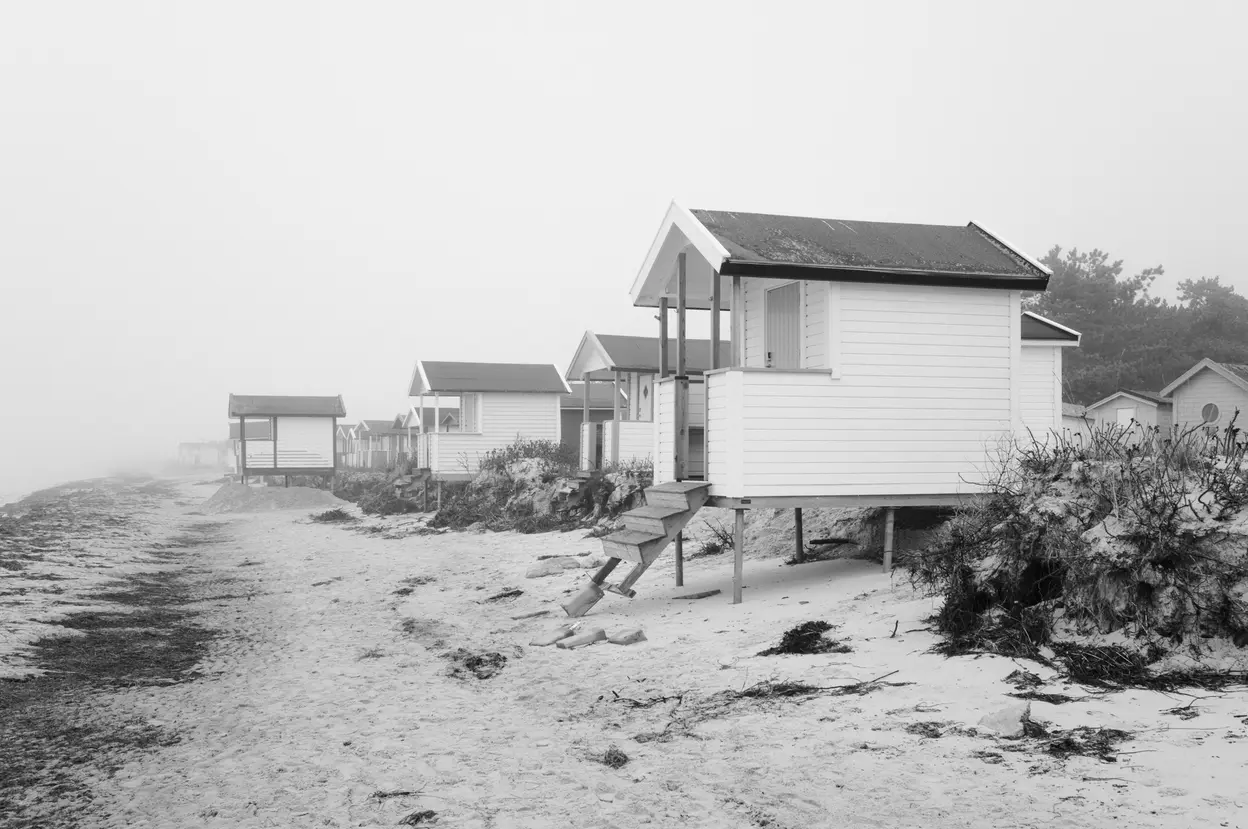 A line of bathing huts fading into fog, the nearest one raised on stilts above cut sand