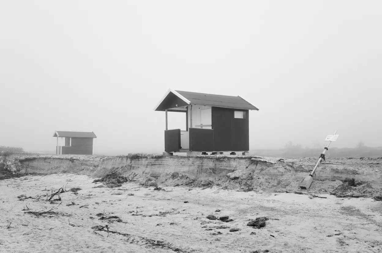 A dark hut and a distant one across flat sand, a sign leaning at the right