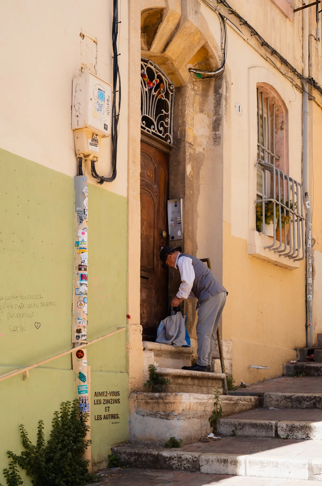 An old man in a waistcoat bending to the door of a yellow house at the top of worn stone steps, a sticker-covered pipe beside him