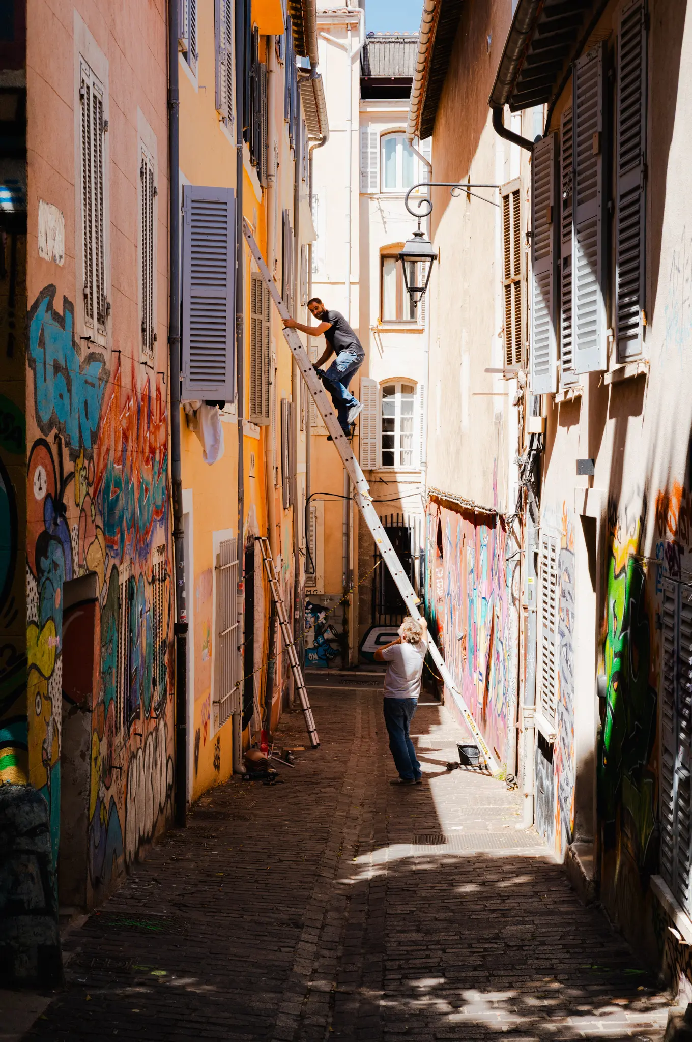 A man up a long ladder against shuttered houses in a narrow graffiti-lined alley, another steadying it from below