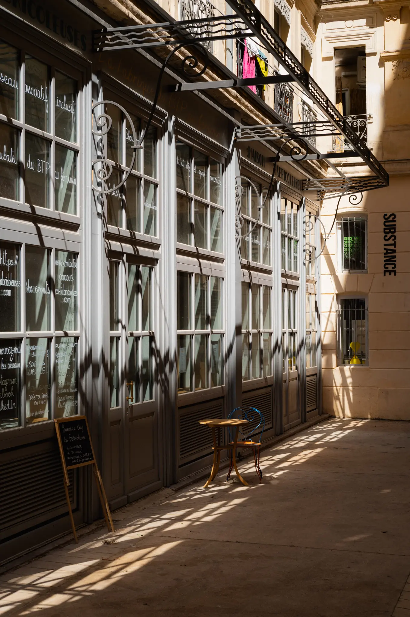 A shaded arcade of tall paned shopfronts, sunlight through an iron canopy striping the paving and a lone café table
