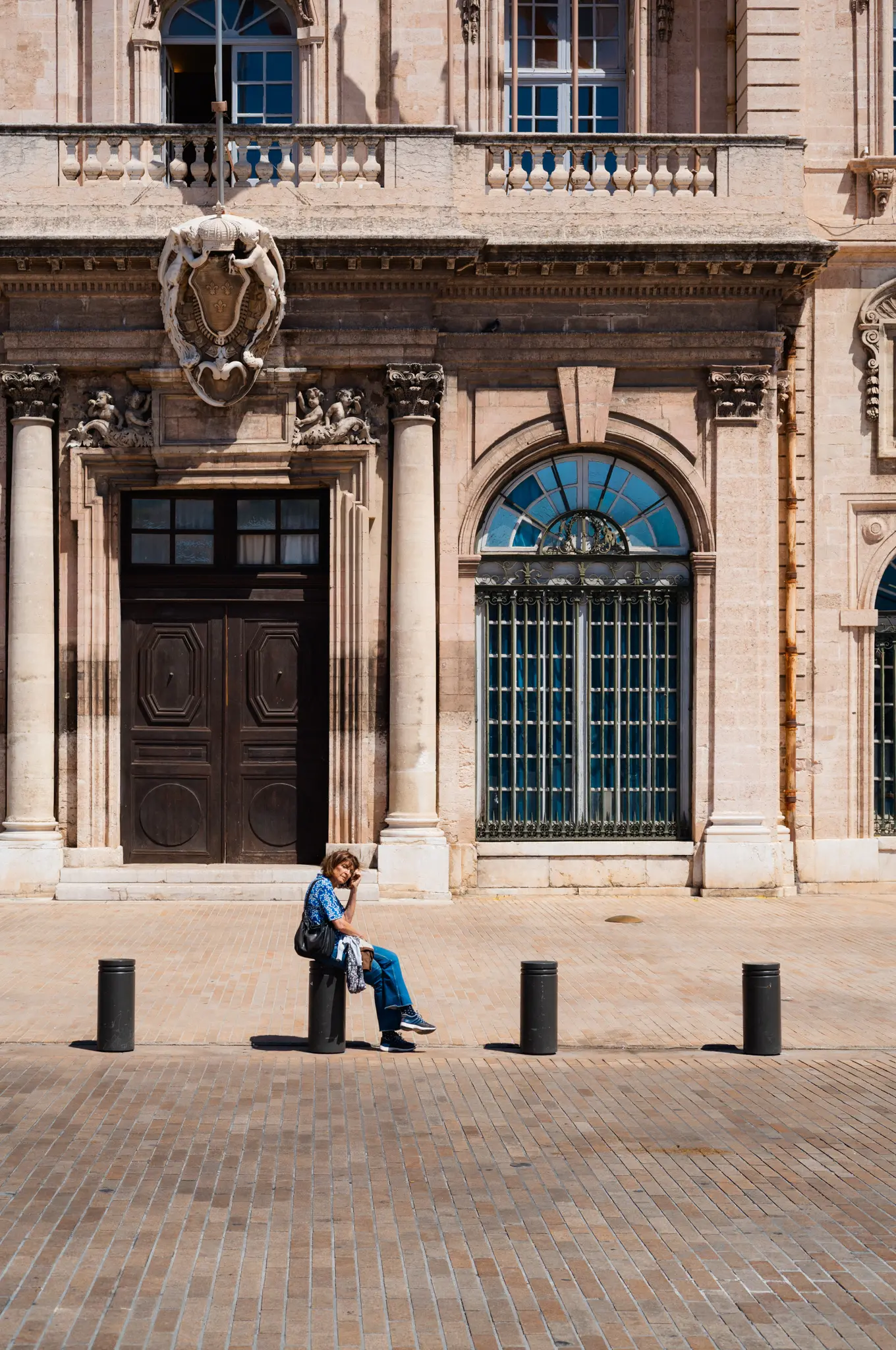 A woman sitting on a bollard in an empty square, the carved stone front of an old building behind her