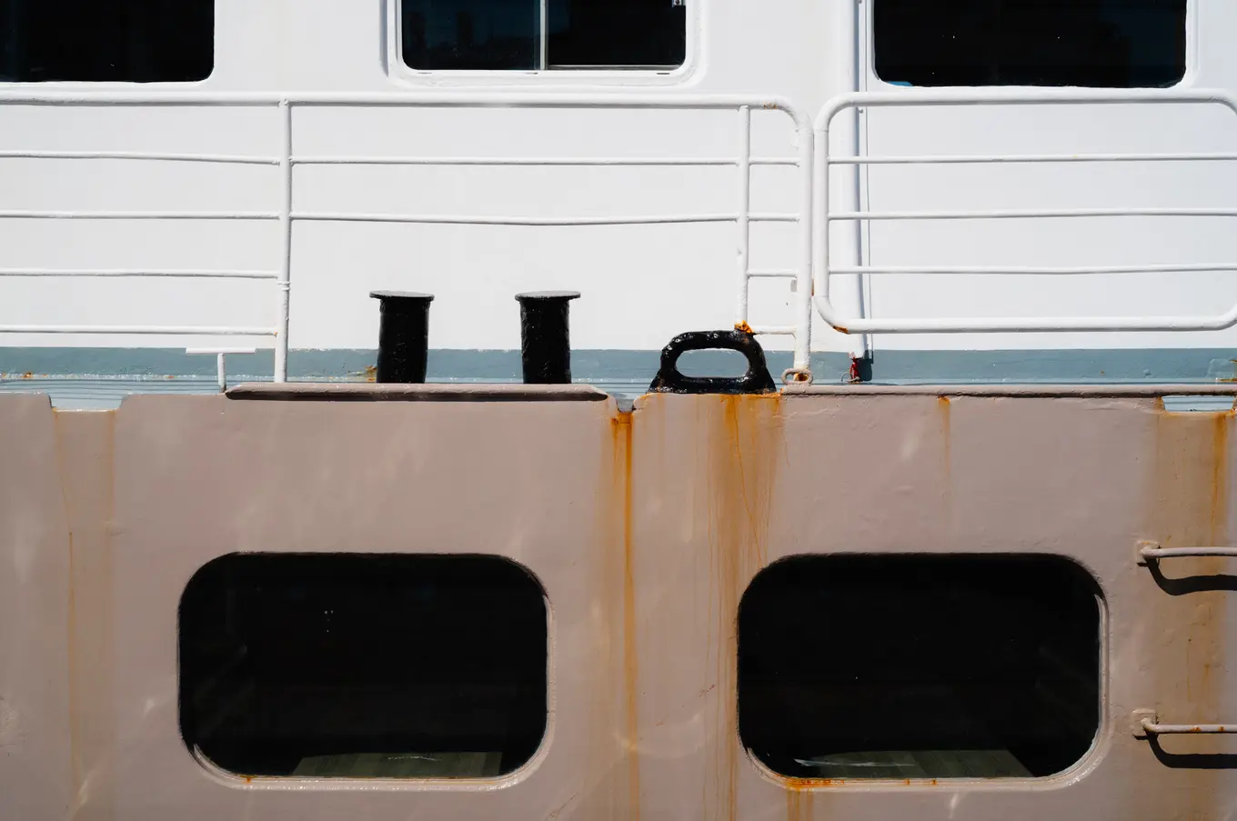 The side of a ship, two dark portholes in a rust-streaked hull beneath white railings and a pair of bollards