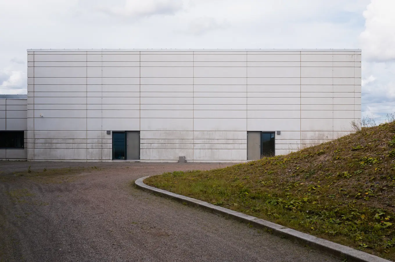 A long white wall with two dark doors, a path curving past a grass mound