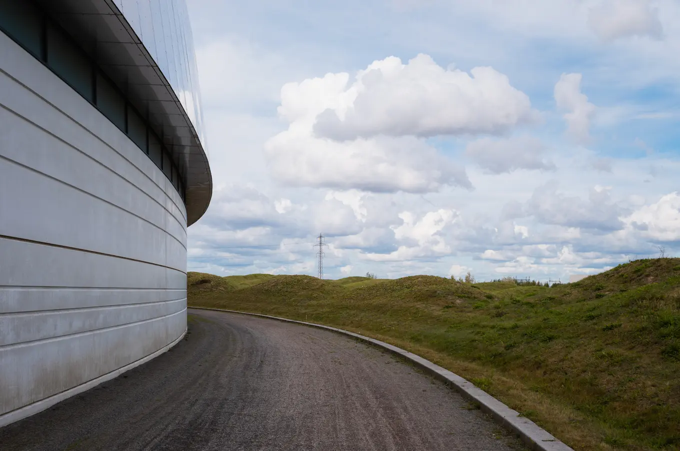 The ring wall curving out of frame beside a road through grassy mounds, a pylon in the distance