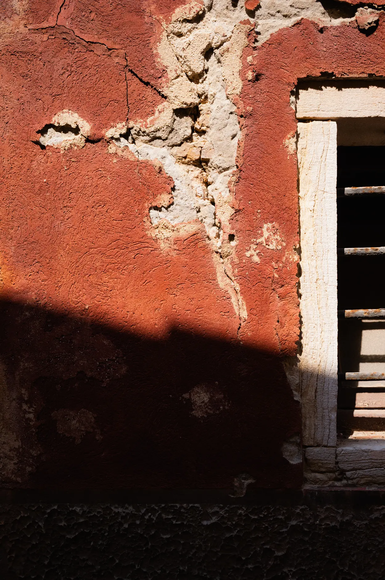 Cracked red render beside a white window frame, half of it in hard shadow