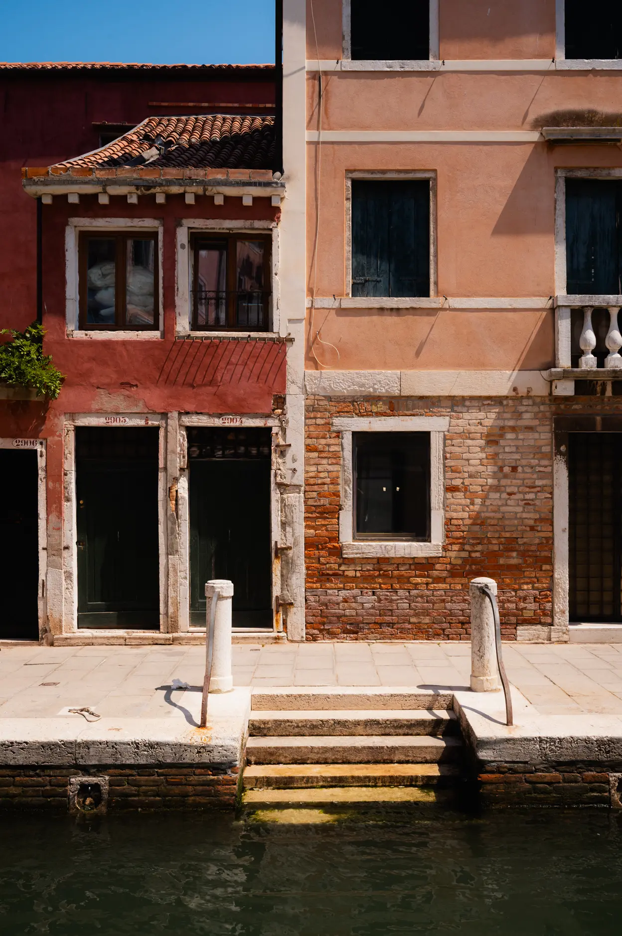 Steps down to a canal between a red and a pink house, two bollards at the water