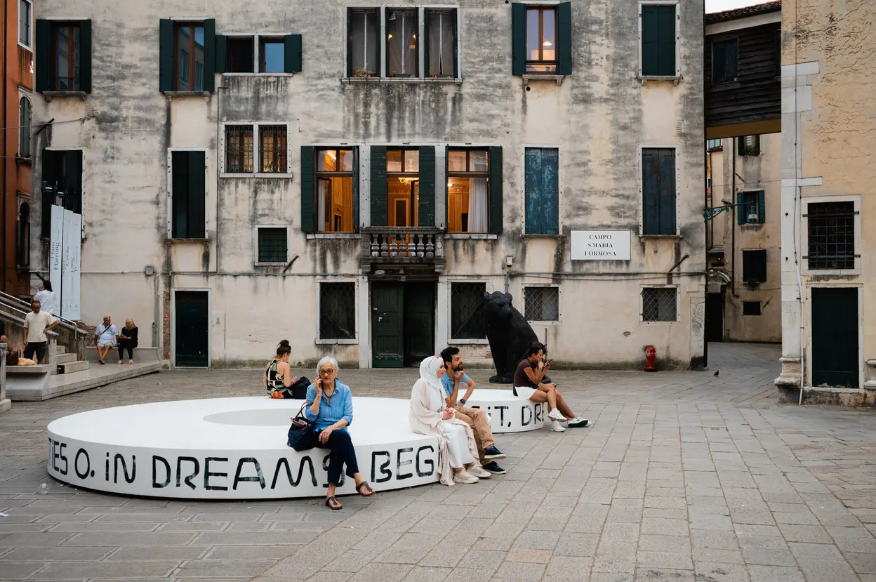 People sitting on a low white sculpture lettered with words, across a campo