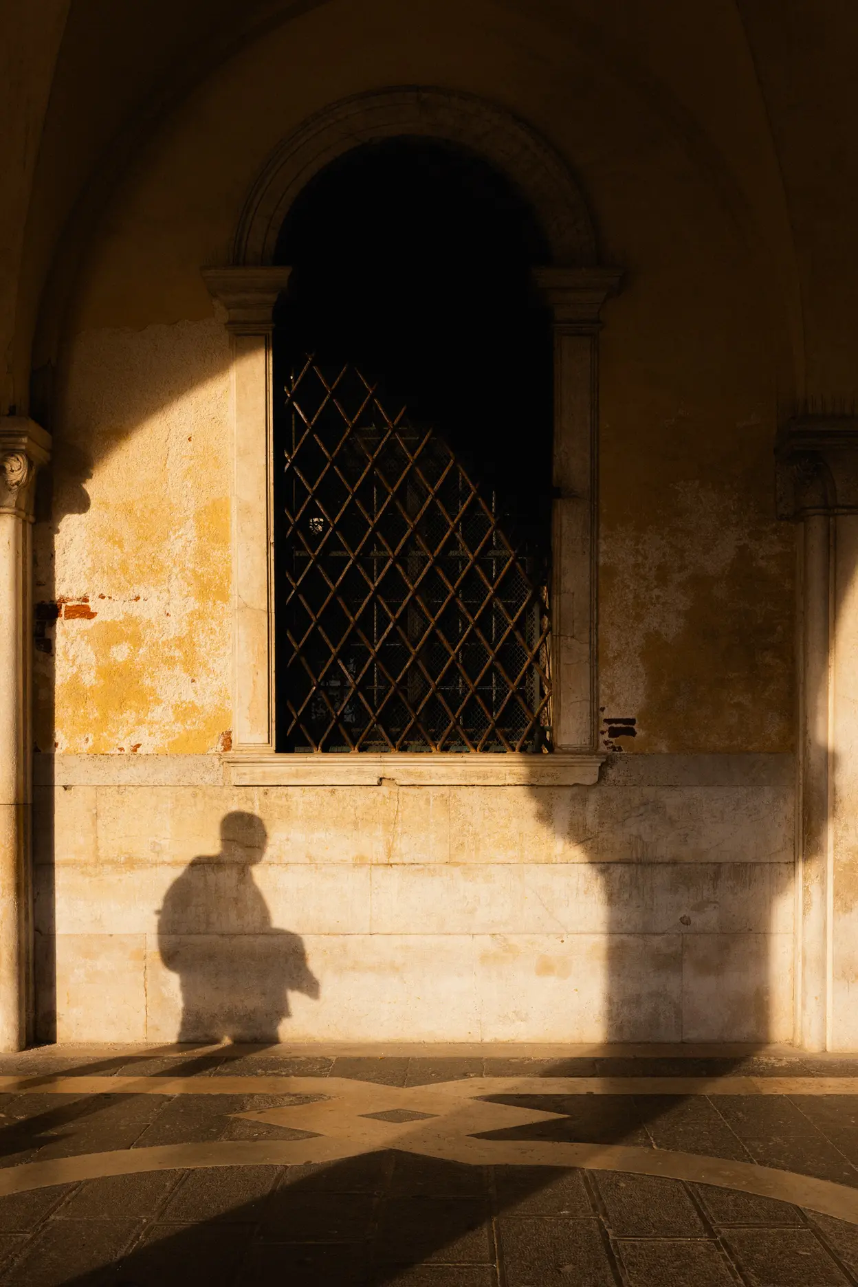A grated window in a yellow wall, the shadow of a figure thrown across the stone