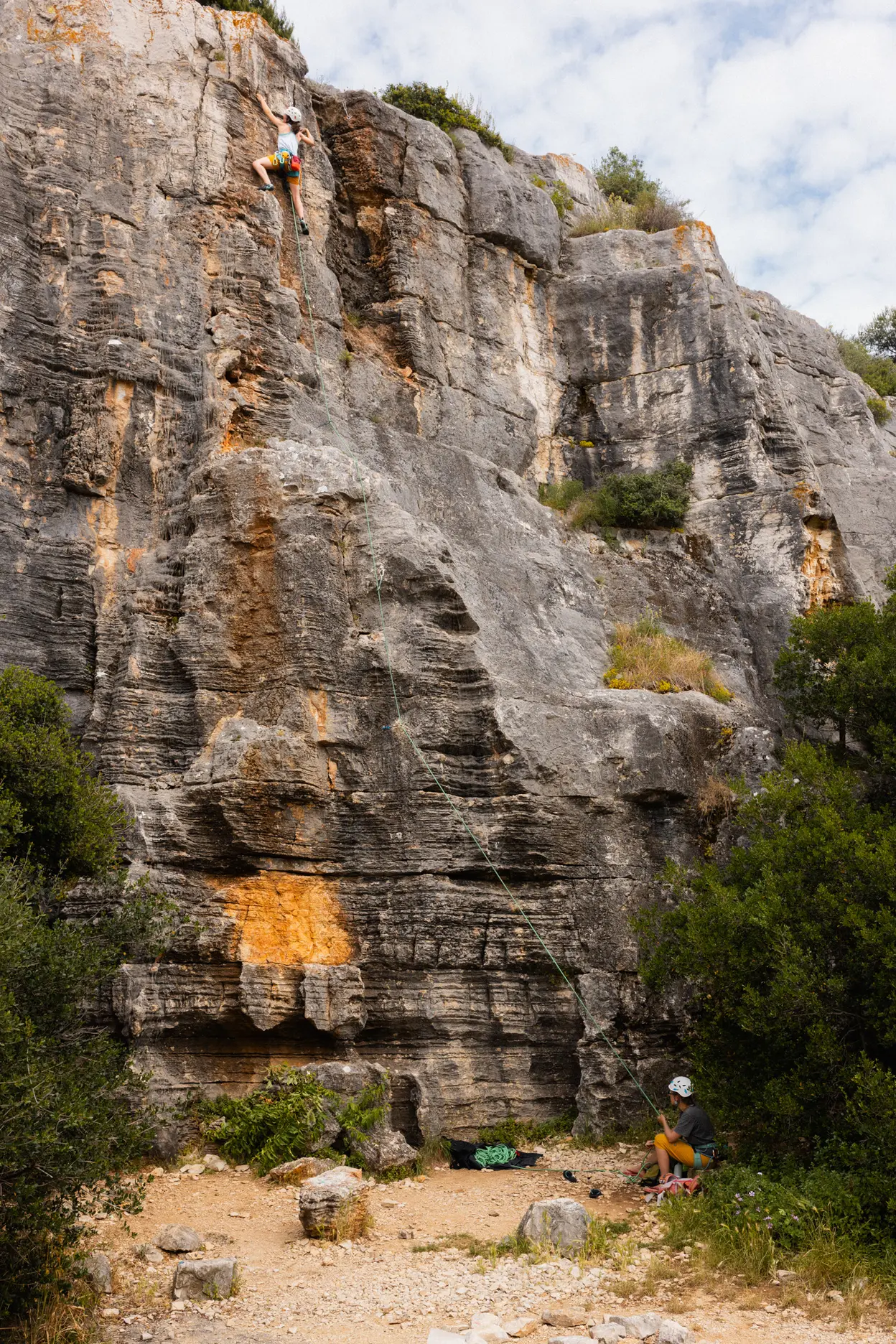 A climber high on a limestone crag, someone belaying at the foot of it