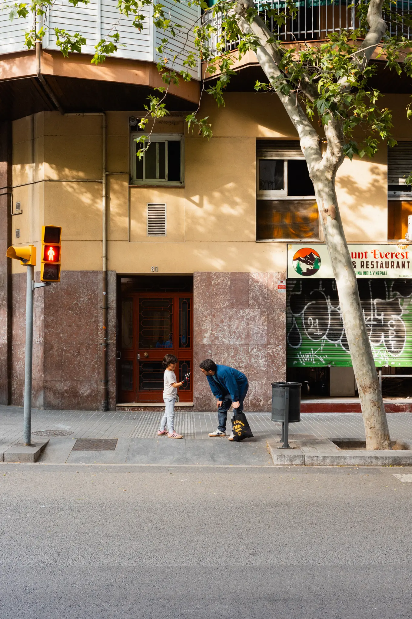 A child waiting at a crossing while another crouches over a scooter, a plane tree filling the corner above them